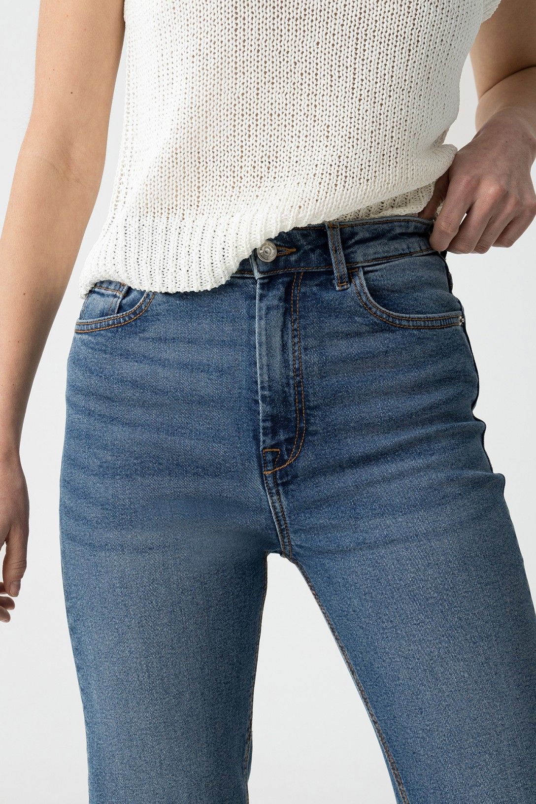 "Dettaglio ravvicinato della vita alta dei jeans Mom_Slim_39 con bottone metallico e zip, indossati con top bianco a maglia"