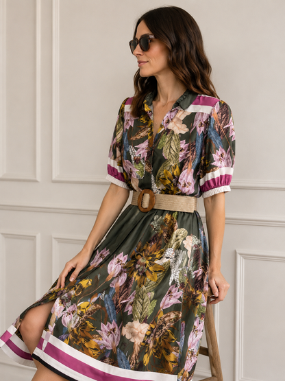 Abito donna botanico militare indossato, vista laterale tre quarti, fantasia con foglie tropicali e fiori esotici su fondo verde scuro, cintura in rafia intrecciata, bordo gonna a righe fucsia, cotone Made in Italy
