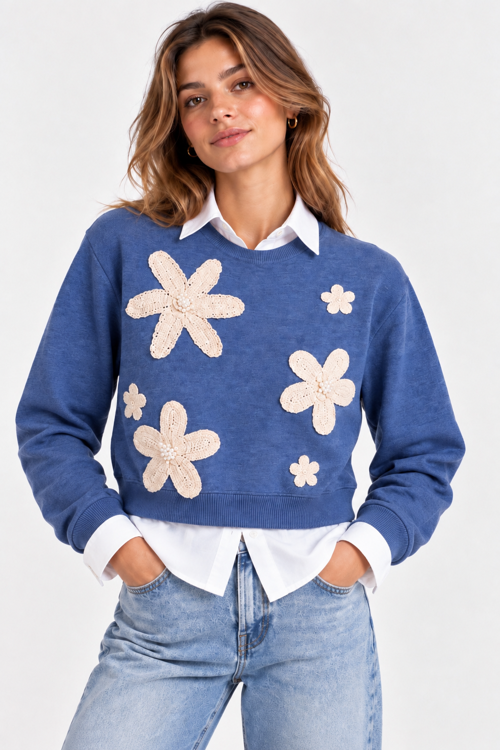 Maglia donna crop azzurra 8090 indossata sopra una camicia bianca, con girocollo, maniche lunghe e applicazioni floreali color panna sul davanti.