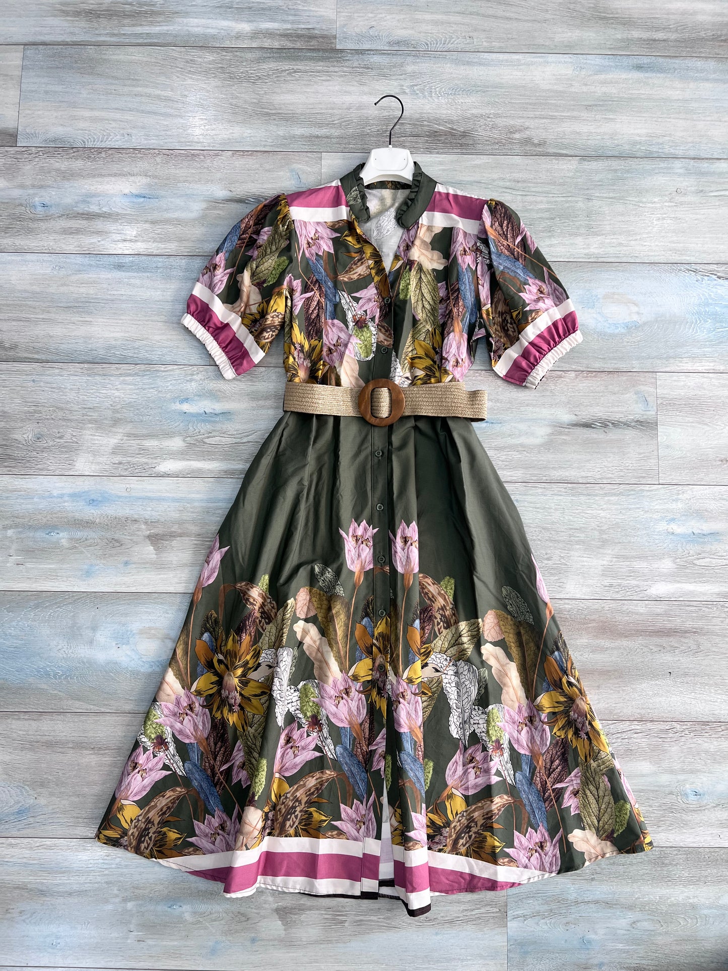 Abito midi donna militare fantasia botanica su gruccia, vista frontale intera, gonna ampia con fiori rosa e foglie tropicali su fondo verde scuro, bordo a righe fucsia, cintura in rafia, cotone Made in Italy
