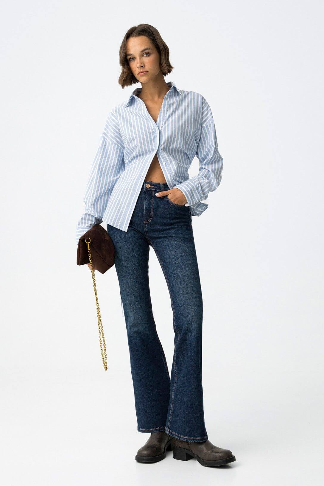 Jeans donna blu denim flare vita alta modello Zoe 64 Frarò, vista frontale su modella con camicia a righe azzurra aperta, borsa a tracolla marrone e stivali Chelsea
