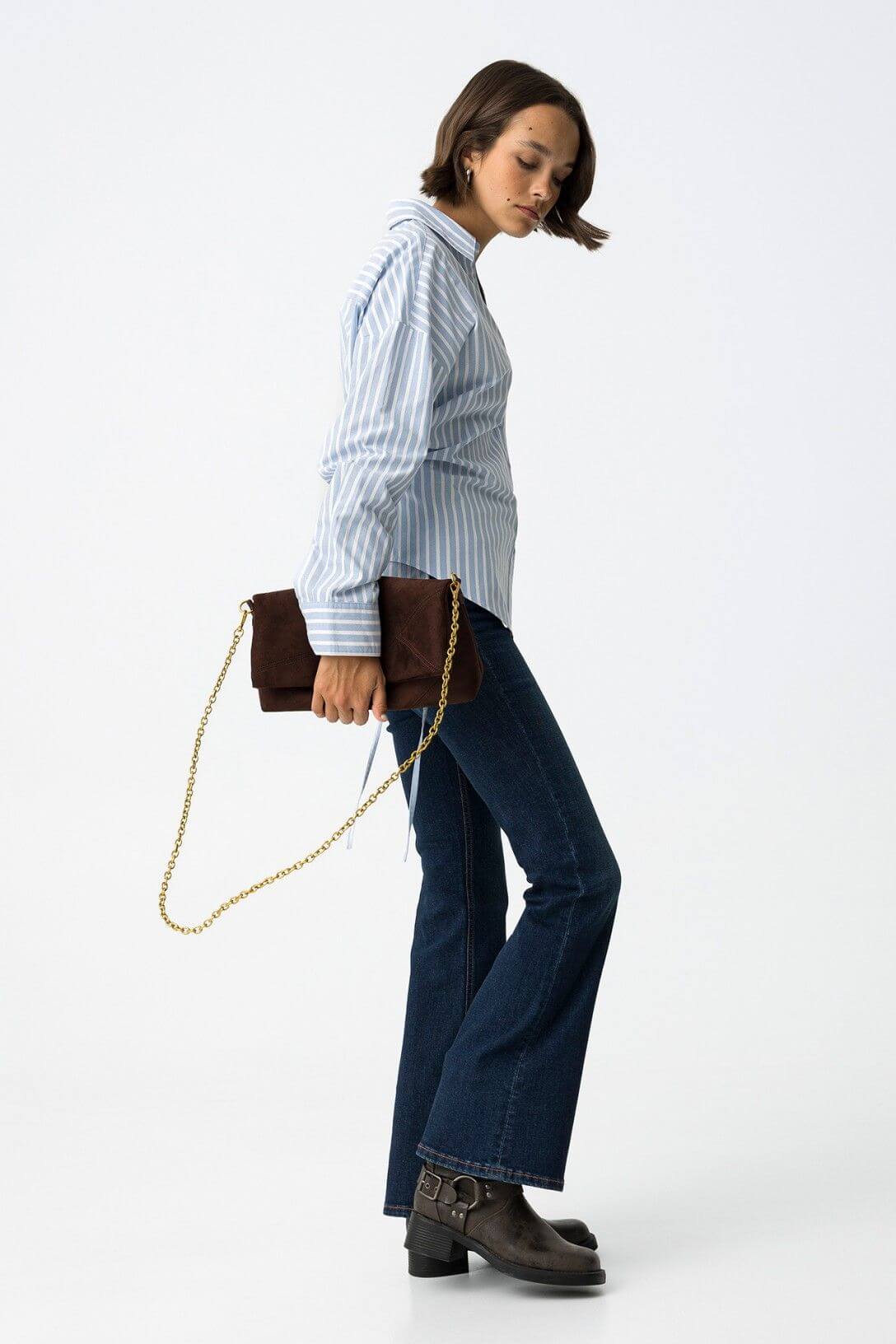 Vista laterale jeans donna blu denim flare vita alta Zoe 64 Frarò, modella con camicia a righe azzurra e borsa clutch marrone con catena dorata, stivali biker neri
